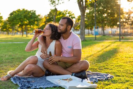 Peaceful sweet couple enjoying dinner in park. Man and woman sitting on grass near box of pizza, drinking beer and hugging. Romantic dinner outdoors conceptの写真素材