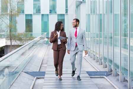 Diverse coworkers going down urban footpath outside. Business man and woman walking near office, using tablet and talking. Conversation outdoors conceptの写真素材