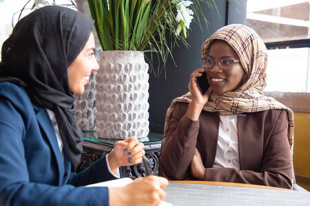 Joyful Muslim businesswoman talking on mobile phone in coffee shop. Her colleague writing notes in...