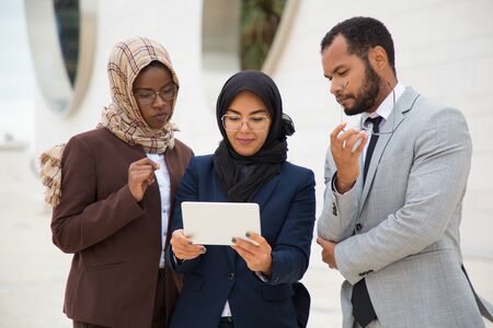 Multiethnic business group watching content on tablet outside. Businessman and Muslim businesswomen standing outdoors and using tablet together. Communication or diversity conceptの写真素材