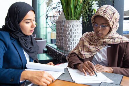Serious female colleagues studying documents in cafe. Young Muslim business women reading and analyzing papers. Paperwork conceptの写真素材