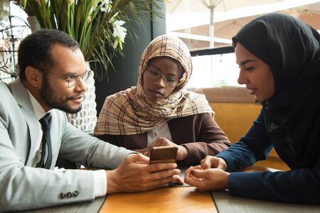 Excited diverse coworkers watching content on smartphone. Businessman and Muslim businesswomen sitting in cafe, man showing phone screen to colleagues. Digital technology conceptの写真素材