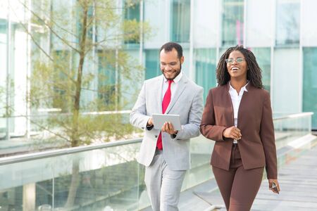 Cheerful coworkers discussing work issues outside. Business man and woman walking near office building, using tablet, talking, smiling, laughing. Walking businesspeople conceptの写真素材