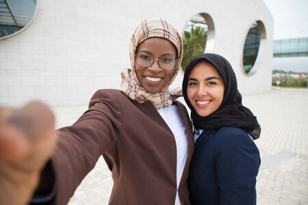 Joyful female corporate friends taking selfie outside. Muslim business women in hijabs holding mobile phone, hugging each other, posing and smiling at screen. Business and friendship conceptの写真素材