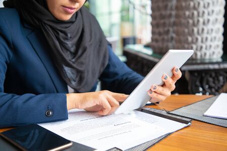 Businesswoman reading contract and consulting internet. Young Muslim business woman sitting in cafe, reading papers and using tablet. Digital communication conceptの写真素材