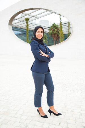 Happy proud Muslim businesswoman posing outside. Full length portrait of young business woman in black hijab and office suit standing for camera with arms folded. Muslim professional conceptの写真素材