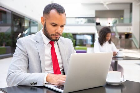 Focused confident entrepreneur working in office lobby. Business man sitting at table, using laptop,...