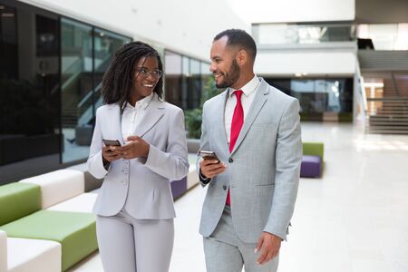 Happy business colleagues using mobile phones and chatting. Business man and woman walking through office hallway, holding smartphones and talking. Corporate communication conceptの写真素材