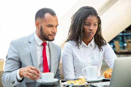 Exited diverse business colleagues watching presentation on laptop. Business man and woman sitting in cafe, drinking coffee, eating, looking at computer screen. Coffee break conceptの写真素材