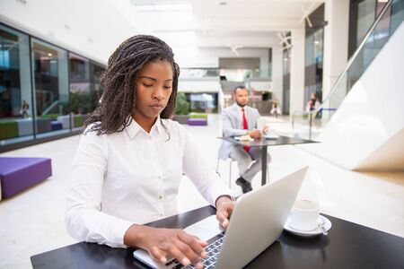 Serious female professional working on project during coffee break n office hall. Young African American business woman using laptop, man drinking coffee in background. Wireless technology conceptの写真素材