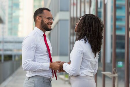 Cheerful multiethnic couple meeting outside during work break. Business man and woman standing in city street, holding hands, smiling and talking. Office dating conceptの写真素材