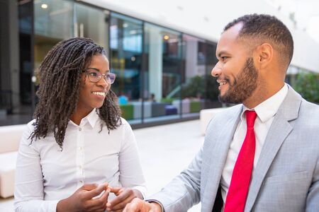 Happy diverse office couple chatting during work break. Business man and woman sitting in office hall, talking and laughing. Corporate relationship conceptの写真素材