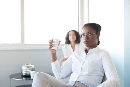 Confident female professional enjoying coffee break in office lounge. Young business woman sitting in armchair, holding disposal cup, drinking coffee and looking away. Coffee break conceptの写真素材