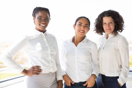 Happy female business team posing together near office window. Businesswomen standing for camera and...