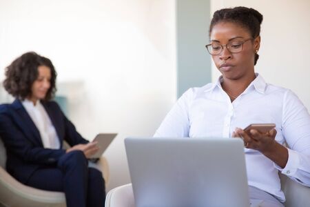 Focused young female manager working in office lounge. Black business woman sitting in armchair, using laptop and smartphone in office lobby. Digital communication conceptの写真素材