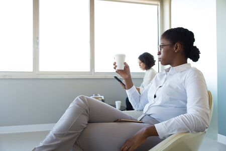 Office employees drinking coffee in office lounge. Young business woman sitting in armchair, holding...