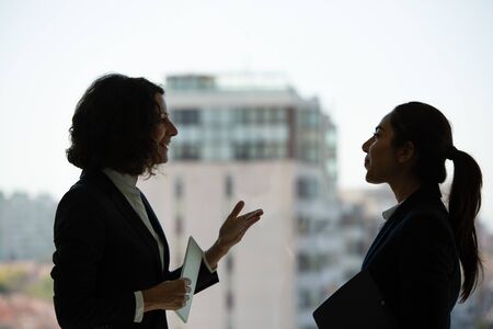 Excited female business colleagues chatting in office corridor. Business women standing near office window and talking. Corporate relationship conceptの写真素材