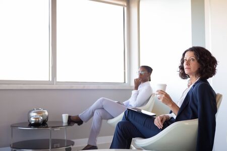 Businesswomen relaxing in office lounge. Diverse business women sitting in armchairs, using tablet,...