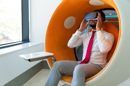Businessman watching virtual presentation. Calm man wearing VR glasses, sitting in interactive chair and adjusting device. Technology or entertainment conceptの写真素材