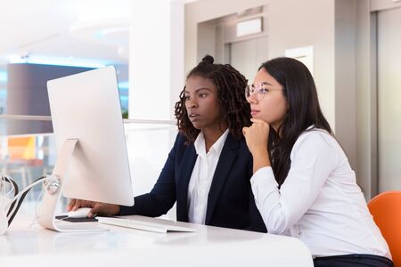 Female office colleagues watching project presentation together. Young business women sitting at table, using computer, looking at monitor. Computer technology conceptの写真素材