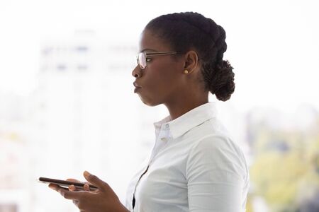 Serious young business lady using mobile phone. Side of African American businesswoman standing against panoramic window, holding smartphone and thinking. Pensive businesswoman conceptの写真素材