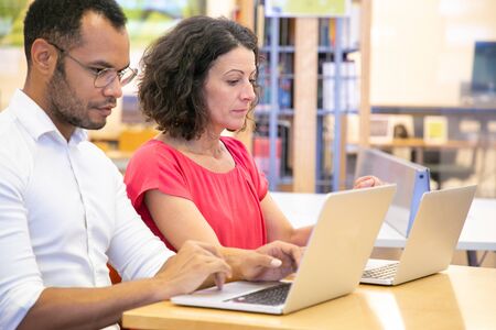 Focused adult students taking tests in classroom. Man and woman in casual sitting at desk, using...