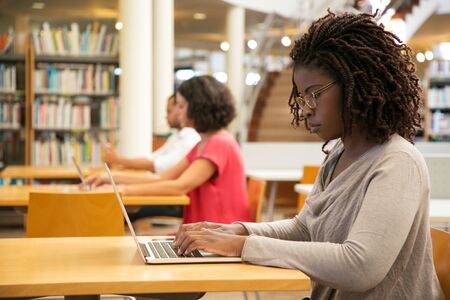 Focused female customer using public wi-fi hotspot in library. Young African American woman sitting at desk and using laptop. Wireless internet connection conceptの写真素材