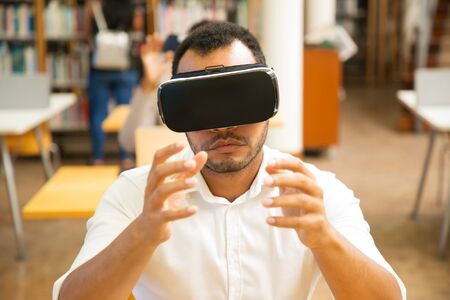 Excited male student using VR simulator during class. Latin man in virtual reality glasses, sitting at desk with laptop and holding air. Simulation conceptの写真素材