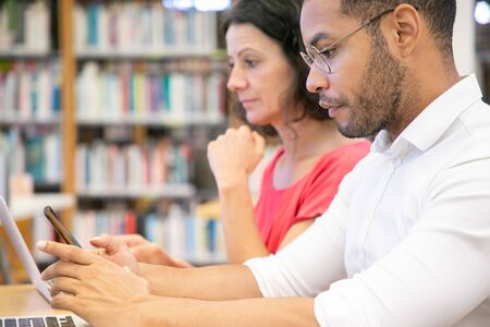 Adult college students using digital devices in library. Man and woman in casual sitting at desk, working on laptop, holding mobile phone, typing message. Digital technology conceptの写真素材