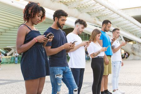 Line of people browsing internet on smartphones outside. Multiethnic men and women standing at city...