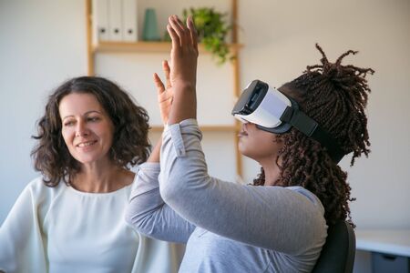 Excited young African American woman wearing VR glasses, touching air. Her female colleague talking...