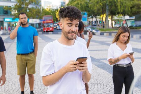 Cheerful guy using smartphone in city street. Young man and woman standing behind him, holding and...