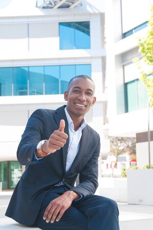 Happy confident business professional making like gesture outside. African American businessman sitting near office building and showing thumb up. Confident business man conceptの写真素材