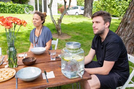People sitting at table in yard. Focused male and female friends sitting at wooden table with meal...