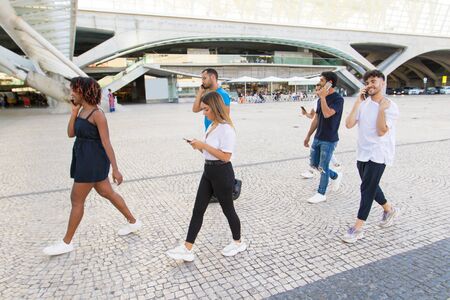Diverse group of people with smartphones walking through city square. Mix raced men and women going...