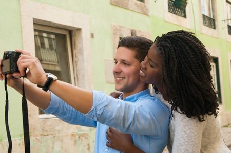 Happy multiethnic couple using photo camera outside. Young man and woman taking self pictures in old town street. Travel and having fun conceptの写真素材