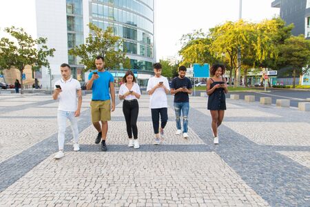 Diverse friends walking together and browsing internet on smartphones outside. Line of men and women...