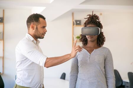 Young African American woman in VR glasses standing in office. Her male colleague instructing her....