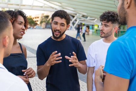 Young mix raced man telling exciting funny story to his friends. Multiethnic men and women standing at city construction, talking, listening. Friendship conceptの写真素材