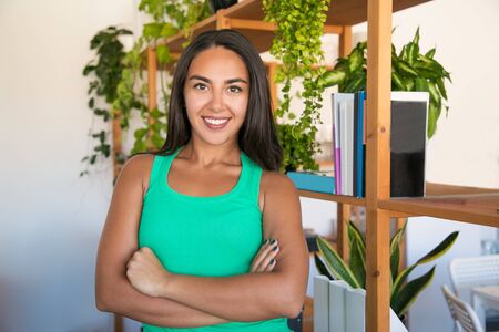 Happy successful female employee in casual posing in modern office. Beautiful young woman with black hair in sleeveless shirt, standing with arms folded and smiling at camera. Female portrait conceptの写真素材