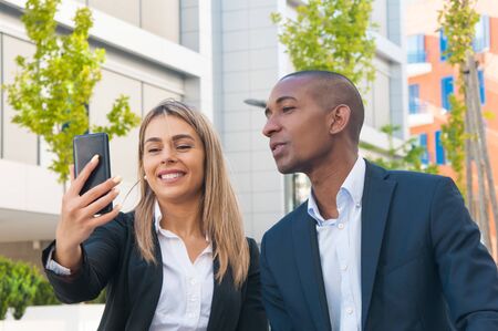 Diverse business colleagues having fun outside. Business man and woman sitting near office building, taking selfie, smiling at phone screen. Selfie conceptの写真素材