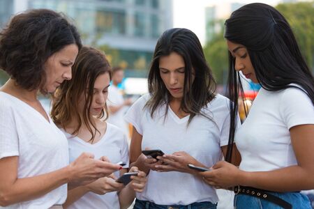Serious friends standing in circle on square with phones. Focused women using smartphones on street. Technology conceptの写真素材