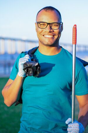Happy male volunteer posing for camera after collecting trash outside. Young man in eyeglasses holding rakes and full plastic bag and smiling. Happy volunteer conceptの写真素材