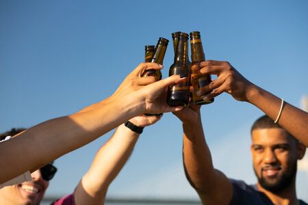 Cheerful friends clinking bottled beer outdoors. Hands of young men and women holding bottles and toasting. Outdoor party conceptの写真素材