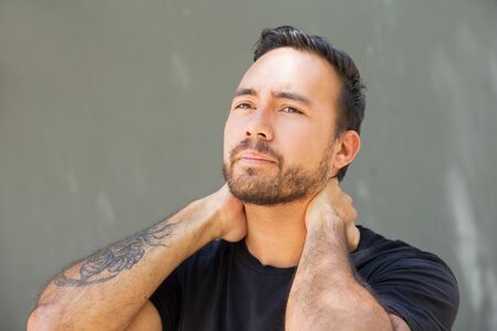 Annoyed guy suffering from neck ache. Young man with tattoo touching neck with both hands. Pain conceptの写真素材