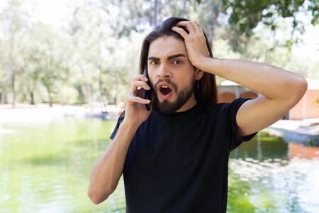 Excited angry long haired guy talking on cellphone outdoors. Handsome bearded young man standing in...