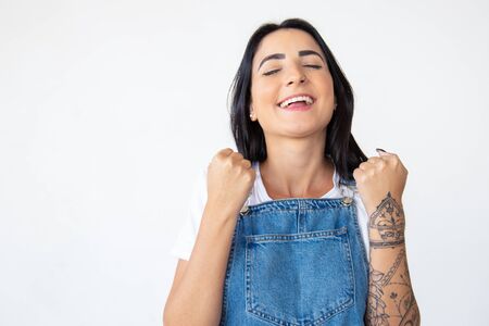 Successful young woman with raised fists. Smiling girl with closed eyes posing on white background. Concept of successの写真素材