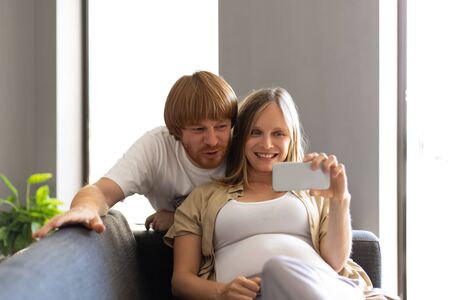 Happy expectant parents using cellphone for selfie or video call. Man bending to pregnant wife lying...