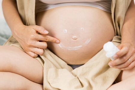 Expectant mother drawing smile with cream on her tummy. Pregnant woman sitting and applying cream to her belly. Pregnancy and body care conceptの写真素材