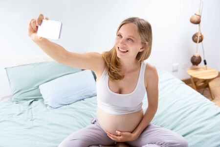 Cheerful expectant mom taking selfie in bedroom. Pregnant woman sitting on bed, using cellphone,...